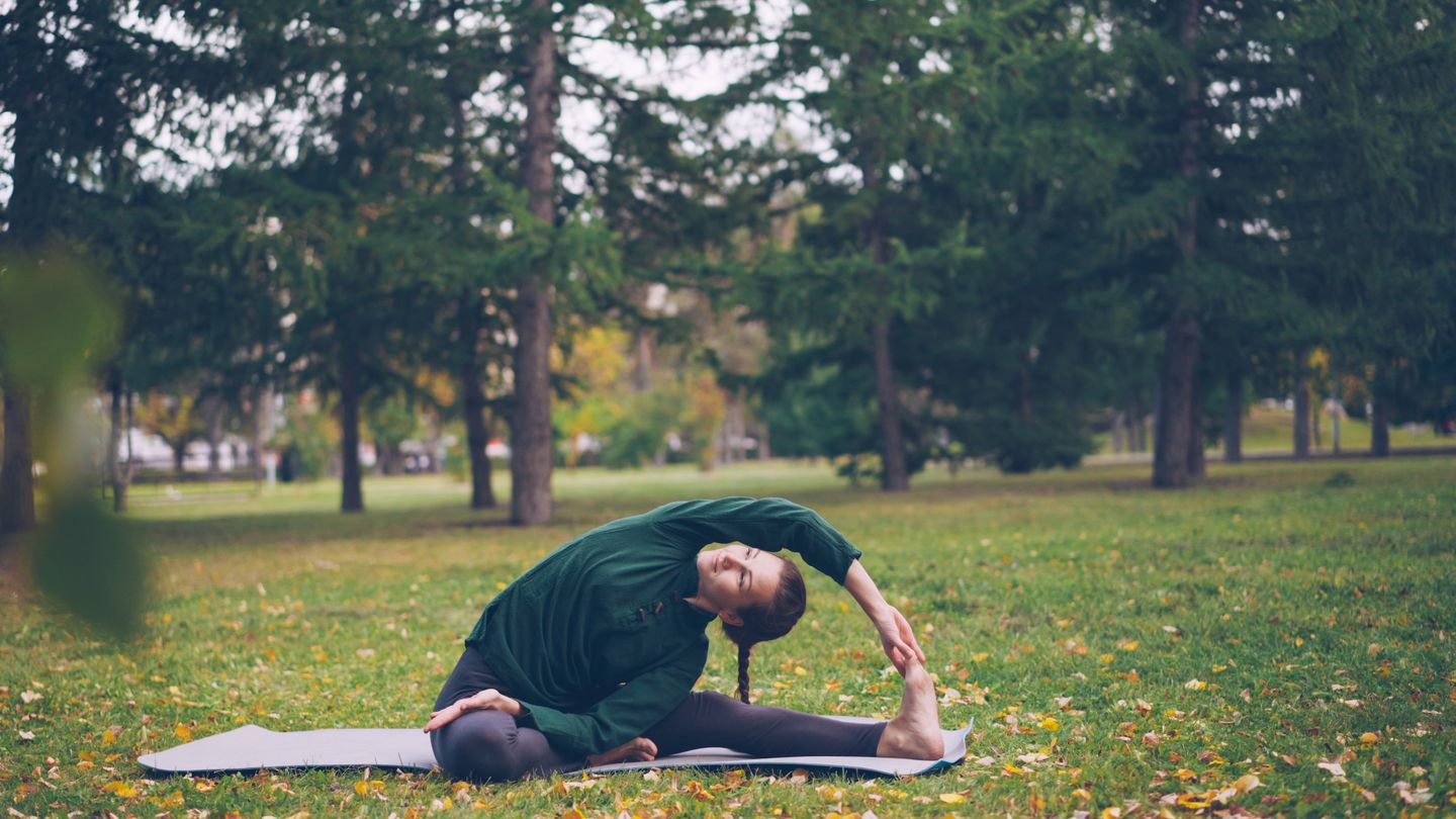 A woman performing a yoga stretch on a mat in a green park setting.